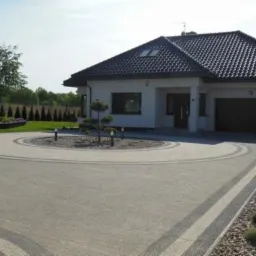 Nowoczesny dom jednorodzinny z ciemną dachówką i jasną elewacją, okrągły podjazd z kostki brukowej w dwóch odcieniach szarości, małe drzewko ozdobne na środku podjazdu, zadbany trawnik i żywopłot...