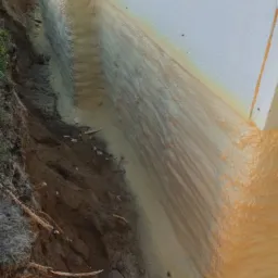 Izolacja fundamentów: Białe płyty izolacyjne z pianką natryskową w wykopie ziemnym, widoczne nierówności terenu i korzenie.