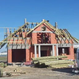 Budowa domu z czerwonej cegły w trakcie wznoszenia więźby dachowej, na pierwszym planie stos drewna budowlanego.