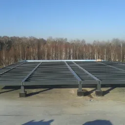 Szara konstrukcja stalowa hali na betonowym fundamencie, widok z góry. W tle drzewa pod błękitnym niebem.