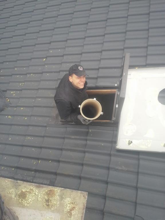 Uśmiechnięty mężczyzna w czapce, wychylony z okna dachowego, trzyma rurę na tle ciemnoszarej dachówki. Praca na wysokości.