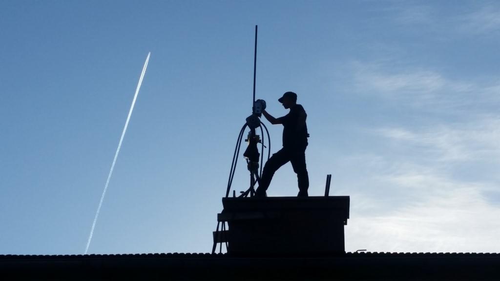 Sylwetka osoby w czapce na dachu, prawdopodobnie podczas pracy przy kominie, na tle błękitnego nieba z widocznym śladem po przelatującym samolocie.