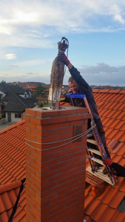 Osoba na drabinie czyści komin z dachu domu z pomarańczową dachówką. Widoczne cegły komina, niebieskie niebo w tle i narzędzia pracy.