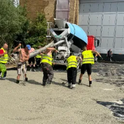 Ekipa w jaskrawych kamizelkach rozlewa świeżą wylewkę z betoniarki na czarną folię. W tle mur z cegły i szara hala.