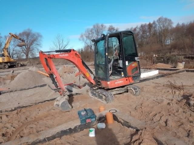 Mini koparka Kubota w wykopie fundamentowym, z widocznym zbrojeniem i elementami instalacji. W tle inna koparka i drzewa.