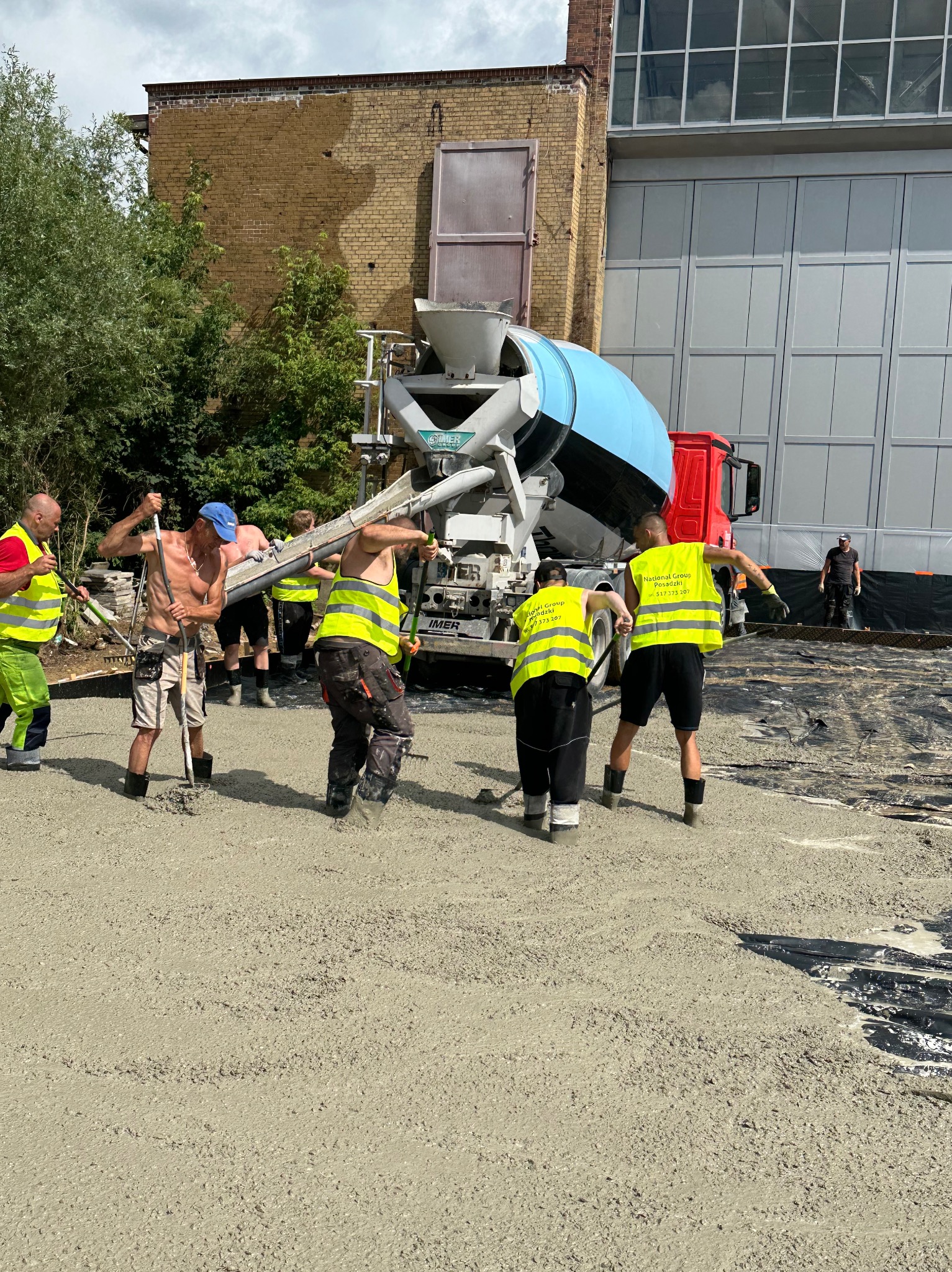 Ekipa w jaskrawych kamizelkach rozlewa świeżą wylewkę z betoniarki na czarną folię. W tle mur z cegły i szara hala.