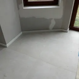 Wykończone wnętrze z jasnymi płytkami podłogowymi, świeżo pomalowanymi ścianami w odcieniach szarości, białymi listwami przypodłogowymi i oknem z drewnianą ramą, widoczne ślady po gładzi na ścianie.