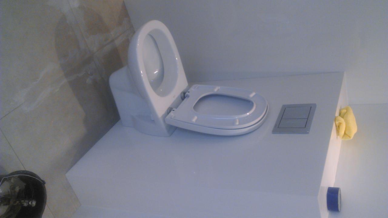 Nowoczesna, podwieszana toaleta z otwartą klapą i szarym przyciskiem spłukiwania, zamontowana na białej zabudowie. W tle beżowe płytki i żółta ściereczka.