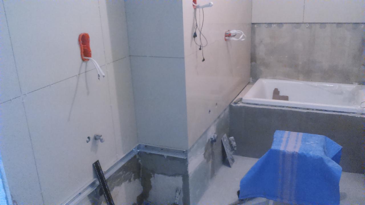 Remont łazienki w trakcie: widoczne płyty karton-gips na ścianach, instalacja elektryczna w puszce, przygotowana wanna. Narzędzia murarskie i poziomica leżą na podłodze.