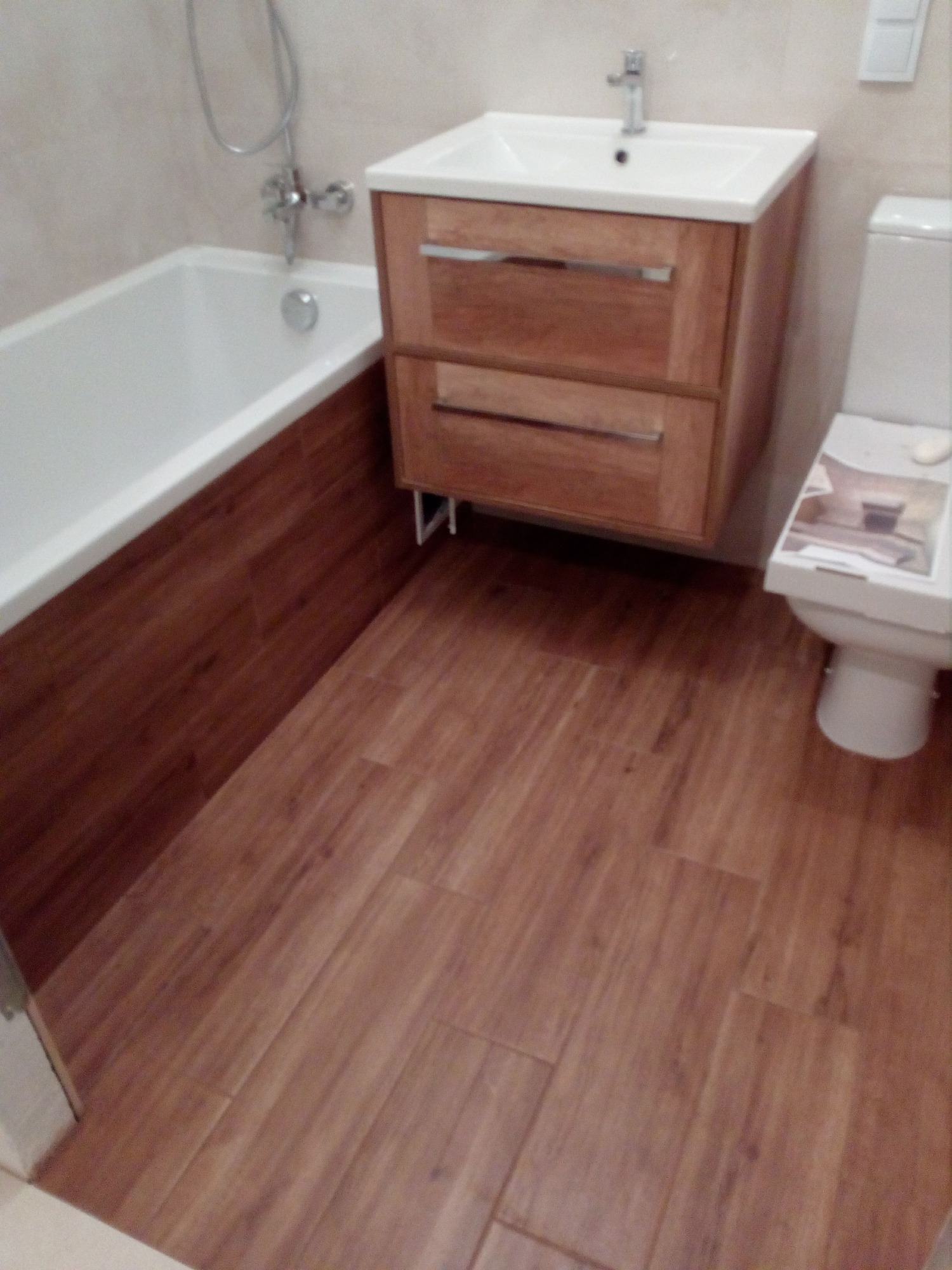 Ukończona łazienka z drewnopodobną podłogą, białą wanną z brązowym obiciem, drewnianą szafką pod umywalką i białą toaletą.