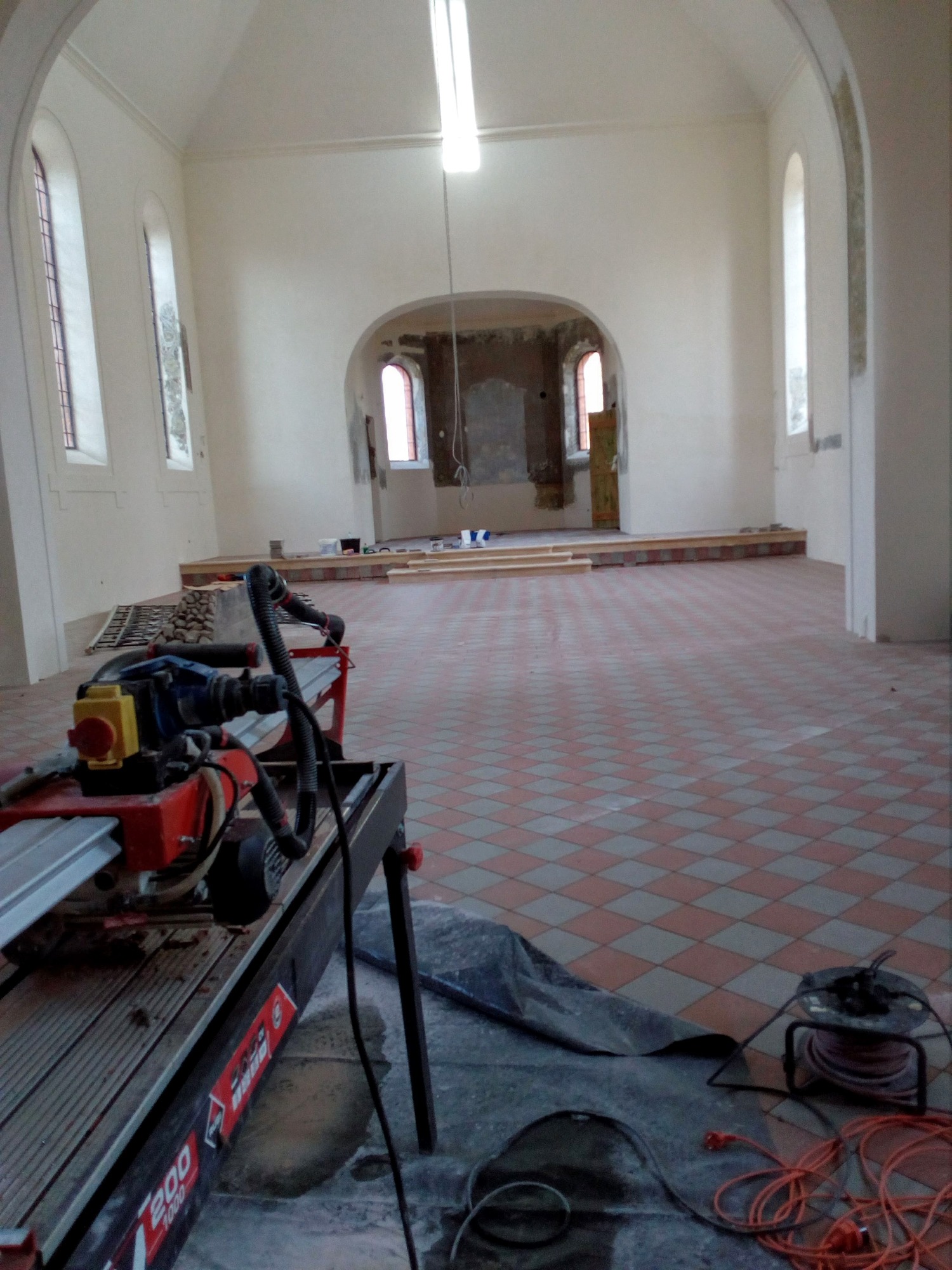Wnętrze kościoła w trakcie remontu, z nową podłogą w kratkę i maszyną do cięcia płytek na pierwszym planie, widoczne okna łukowe i przygotowane podesty.
