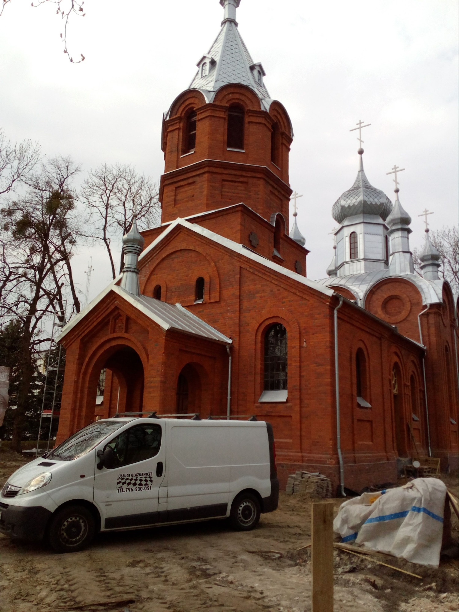 Ceglany kościół z srebrnymi kopułami w pochmurny dzień, z zaparkowanym białym vanem firmy glazurniczej z boku.