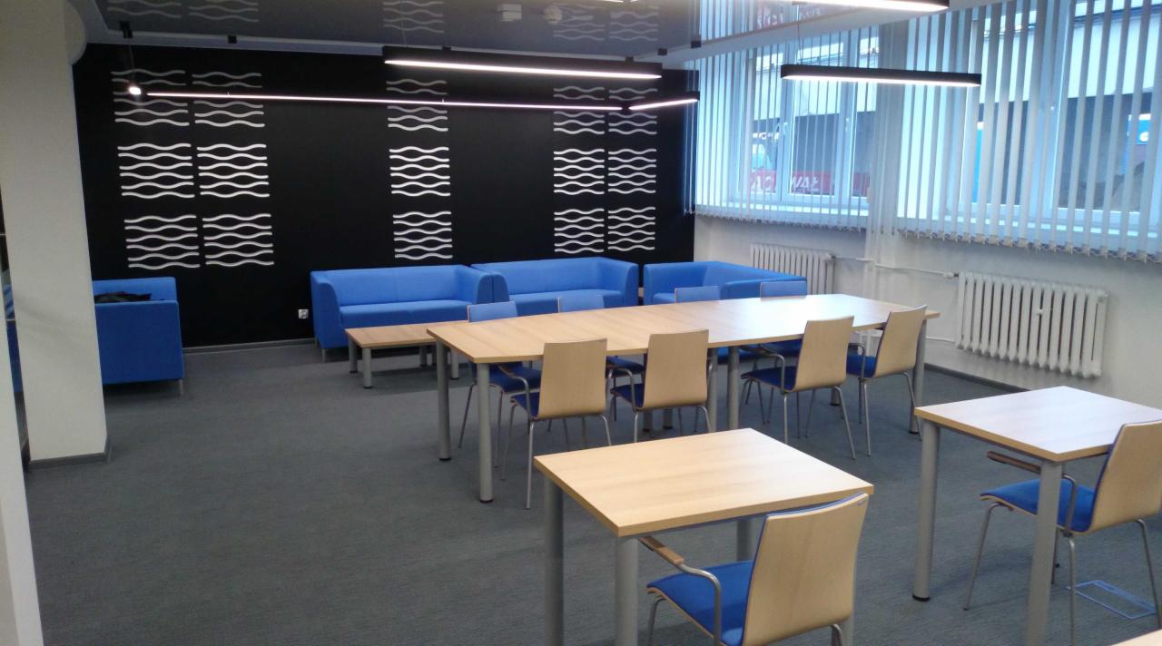 Nowoczesne wnętrze biurowe z niebieskimi sofami, jasnymi stołami i krzesłami. Czarna ściana z białym, falistym wzorem. Białe żaluzje w oknach.