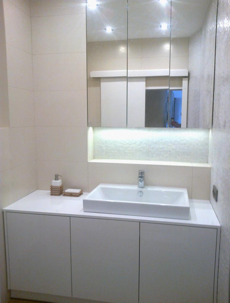 Nowoczesna łazienka: biała szafka pod umywalką z kwadratową umywalką, lustro z oświetleniem LED i beżowe płytki. Widoczne akcesoria łazienkowe.