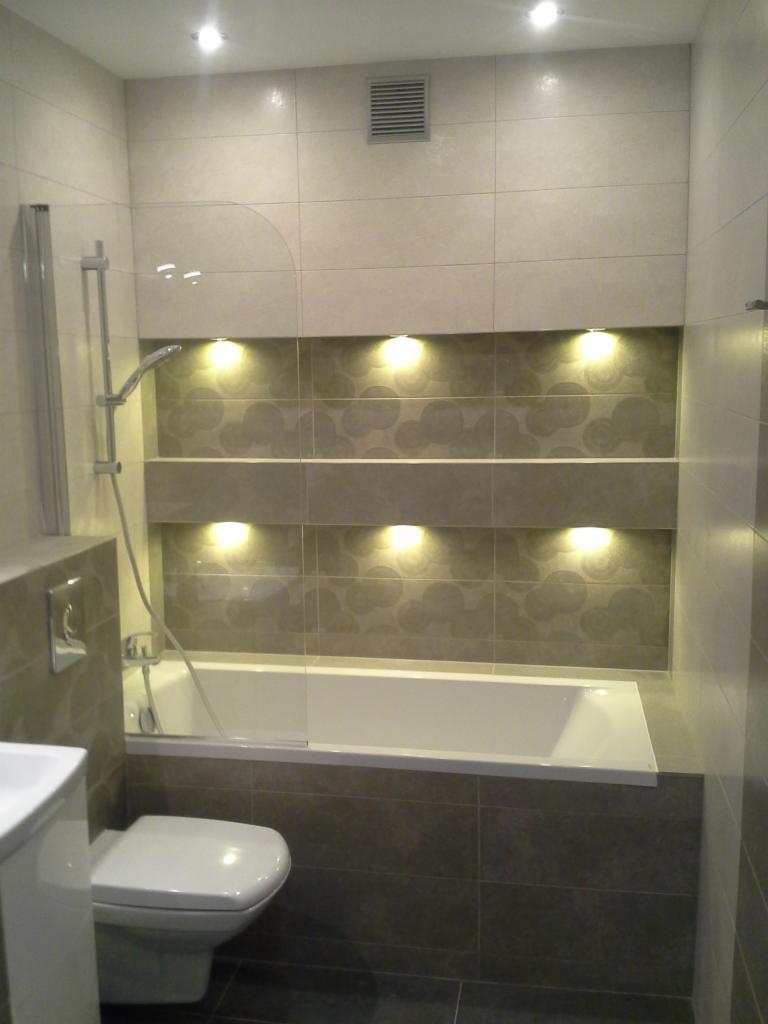 Nowoczesna łazienka z wanną, jasnymi płytkami i oświetleniem wnękowym. Widoczny prysznic i toaleta. Szare płytki kontrastują z bielą wanny.