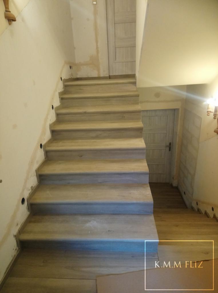 Schody wyłożone płytkami imitującymi drewno, widoczne niedokończone prace wykończeniowe na ścianach i podłodze, w tle drzwi.