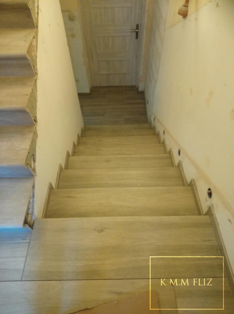 Wykończone schody z płytek imitujących drewno, widok z góry, z widocznymi punktami oświetleniowymi na ścianie i częściowo wykończonymi krawędziami po lewej stronie.