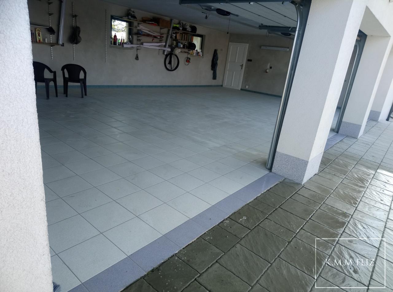 Wykończona podłoga garażu z jasnych, kwadratowych płytek, z szarą listwą przypodłogową i widoczną kostką brukową na zewnątrz, w tle widoczne wyposażenie garażu.