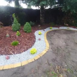 Aranżacja rabaty w ogrodzie z korą, kamieniami i niskimi roślinami, obrzeże z żółtej kostki, widok na trawnik i dom.