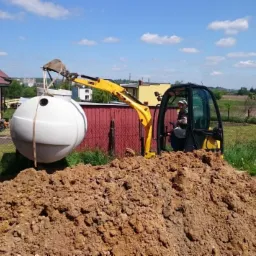 Żółta mini koparka podnosi biały zbiornik na tle domu i zielonego krajobrazu. Widoczny wykop i studzienka rewizyjna.