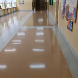 Błyszcząca, brązowo-beżowa podłoga w korytarzu szkolnym, z białym pasem na środku, odbijająca światło z okien i lamp, ściany w kolorze kremowym z tablicami ogłoszeń.