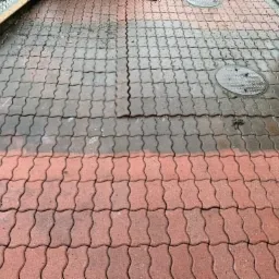 Częściowo umyta czerwona kostka brukowa typu fala, widoczne dwie studzienki kanalizacyjne, różnica w kolorze i zabrudzeniu między czystą i brudną powierzchnią.