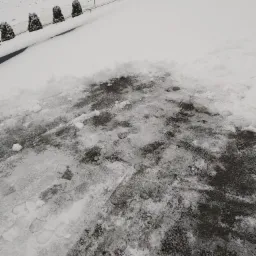 Częściowo odśnieżony plac z widocznymi śladami st&oacute;p, w tle droga z czerwono-białą ciężar&oacute;wką i zasypane śniegiem pole