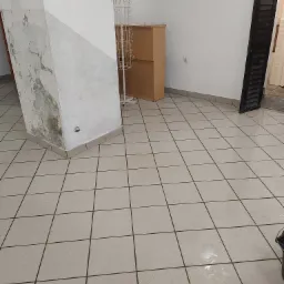 Wnętrze pomieszczenia z jasną, kwadratową podłogą z płytek, kolumną z uszkodzeniami, metalowym stojakiem na ulotki i mopem do podłogi, w trakcie sprzątania widoczne mokre plamy na podłodze.