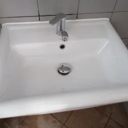 Biała, prostokątna umywalka z chromowaną baterią i korkiem, zamontowana w łazience z szarymi kafelkami.