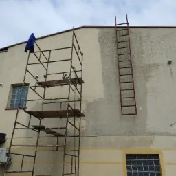 Fragment elewacji budynku z widocznym rusztowaniem, drabina oparta o ścianę i ślady po myciu elewacji, sugerujące prace porządkowe.