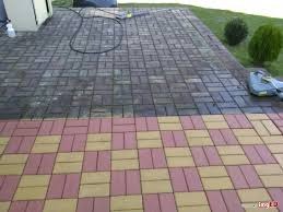 Częściowo wyczyszczona kostka brukowa w dwóch kolorach (różowy i żółty) z widoczną różnicą przed i po czyszczeniu. Na górze widoczny wąż ciśnieniowy oraz butelka z detergentem.