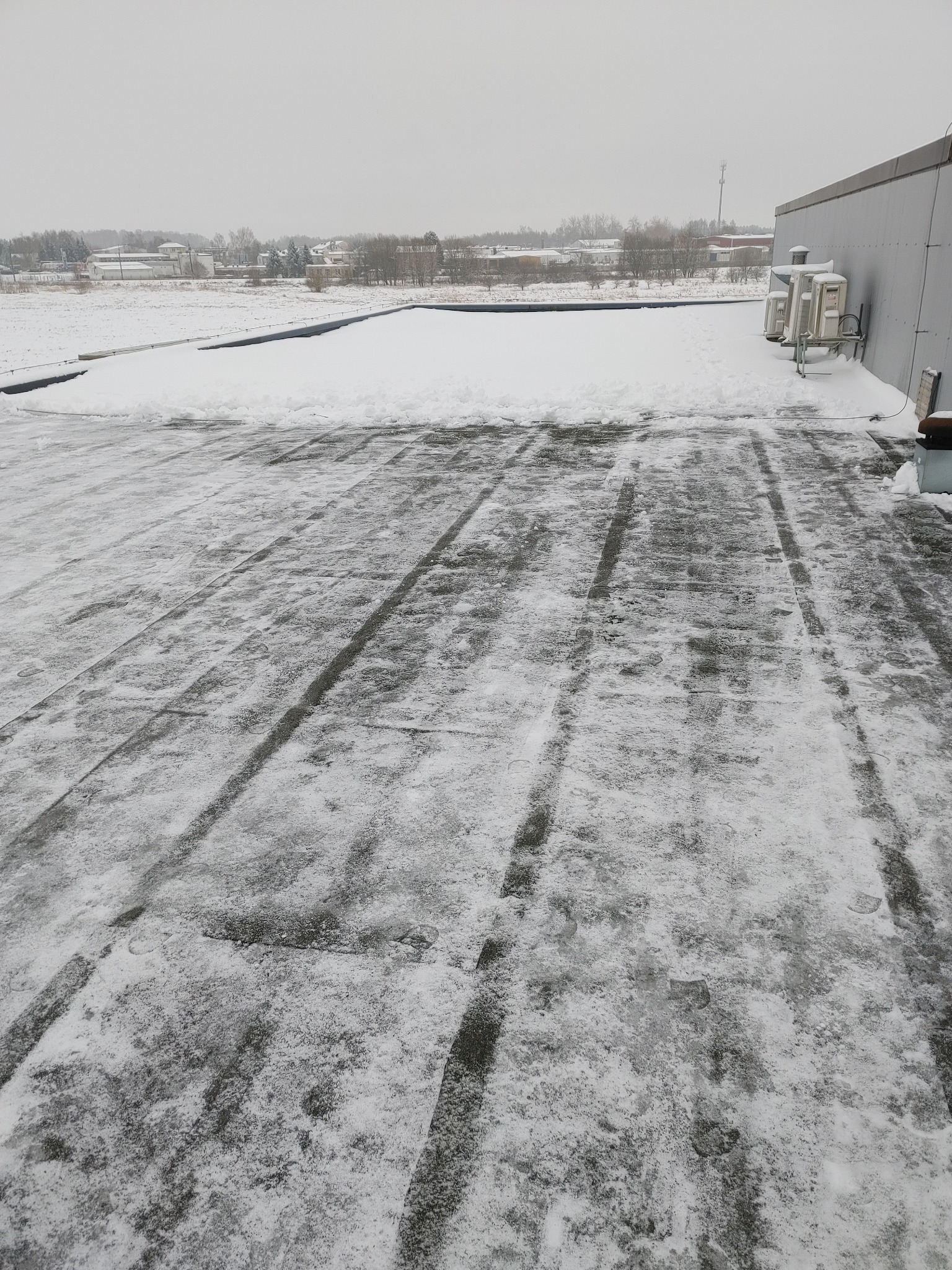 Śnieg na płaskim dachu z widocznymi śladami odśnieżania, w tle zabudowania i pochmurne niebo.