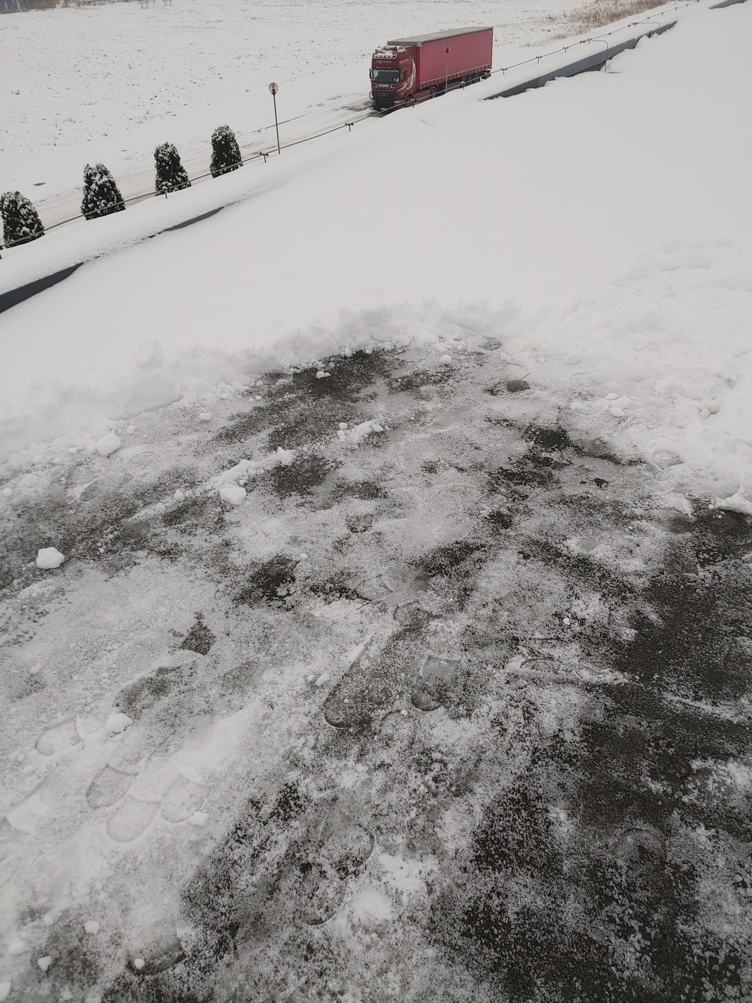 Częściowo odśnieżony plac z widocznymi śladami stóp, w tle droga z czerwono-białą ciężarówką i zasypane śniegiem pole