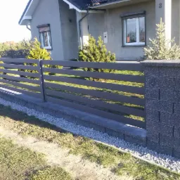 Nowoczesne ogrodzenie panelowe w kolorze grafitowym, z podmur&oacute;wką i słupkami z szarej kostki łupanej, na tle domu z jasną elewacją i zielonym trawnikiem.