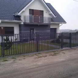 Nowoczesne ogrodzenie z pionowych sztachet w kolorze grafitowym, dopasowane do architektury domu z balkonem z balustradą ze szkła.