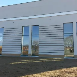 Elewacja budynku z szeregiem wysokich okien i panelami dekoracyjnymi; odbicia w szybach ukazują otoczenie.