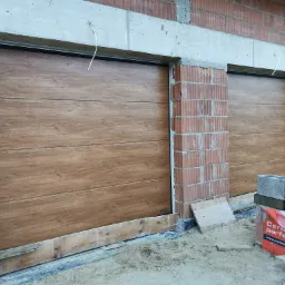 Dwa segmentowe garażowe drzwi z imitacją drewna, zamontowane w surowym stanie budynku z czerwonej cegły, widoczny betonowy nadproże i materiały budowlane na ziemi.