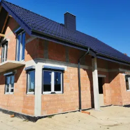 Surowy budynek z cegły z zamontowanymi oknami z roletami zewnętrznymi, ciemna dachówka i błękitne niebo w tle.