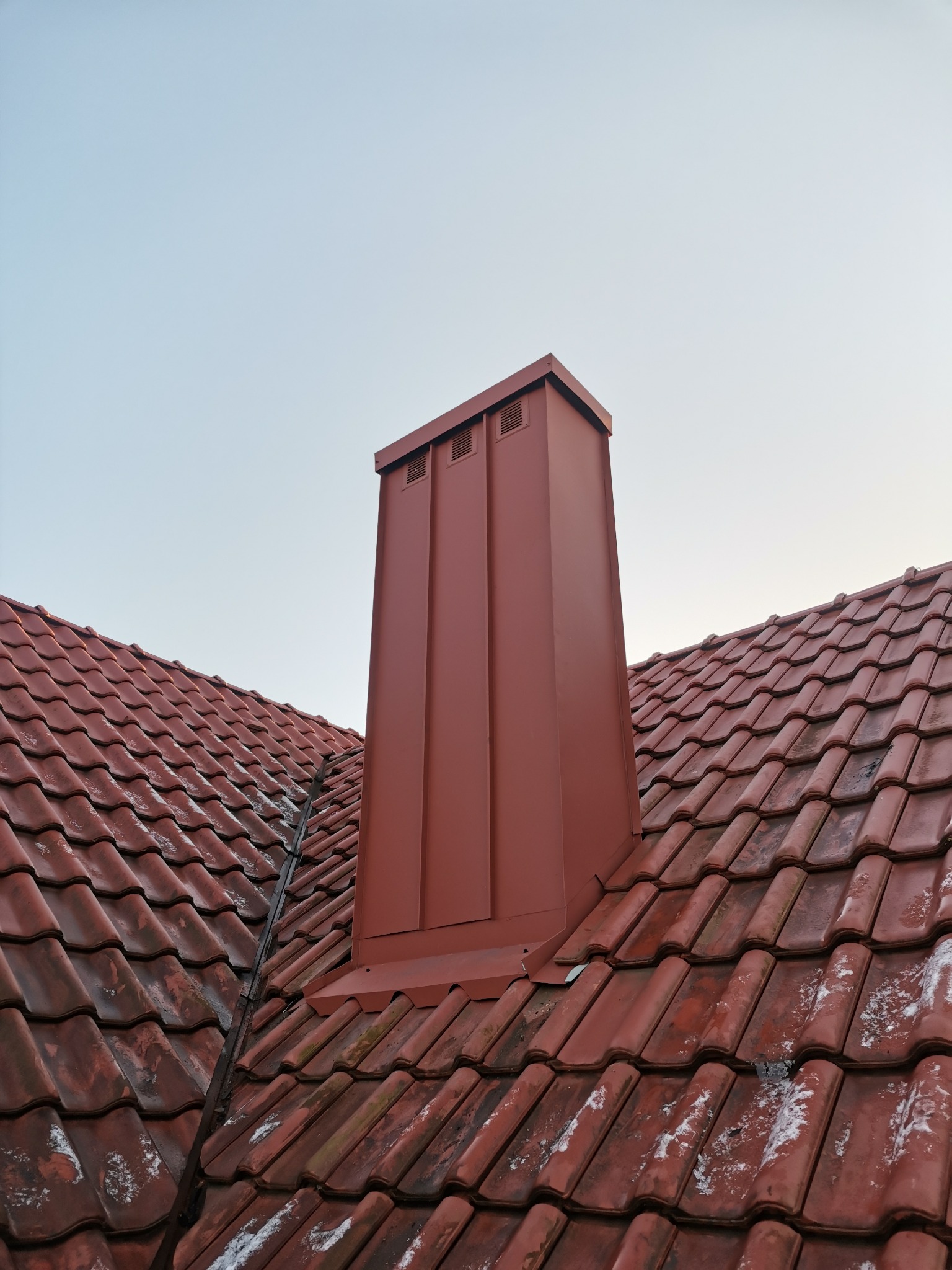 Czerwony, metalowy komin wentylacyjny na dachu z ceramicznej dachówki karpiówki, częściowo pokrytej śniegiem, widok z dołu pod kątem.