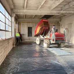 Wnętrze hali w trakcie budowy, na czarnej folii rozłożono zbrojenie, a betoniarka z czerwoną gruszką wjeżdża na teren w asyście dwóch pracowników w odblaskowych kamizelkach.