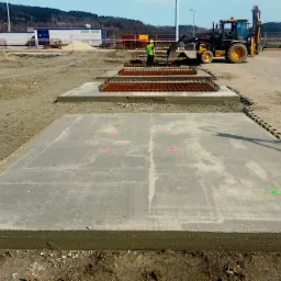 Wylane betonowe płyty fundamentowe na placu budowy, widoczny pracownik w kamizelce odblaskowej i koparka w tle, teren wyrównany, widać zbrojenie pod kolejne płyty, geodezyjne punkty pomiarowe.