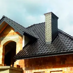 Surowy stan domu z cegły z czarną dachówką i kominem z cegły w trakcie budowy, widoczny łuk okienny.