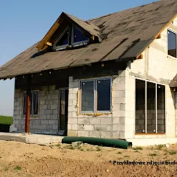Niedokończony dom z białego bloczka, z widocznym zadaszeniem werandy wspartym na drewnianych słupach i oknami wstawionymi w surowy mur, na tle zielonego pola i błękitnego nieba.