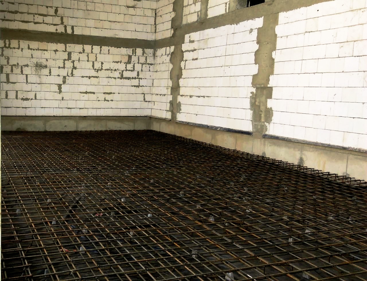 Siatka zbrojeniowa ułożona na podłożu wewnątrz budynku z murem z bloczków betonowych, przygotowana do wylewki fundamentu.