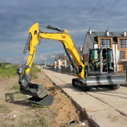 Żółta koparka na gąsienicach z łyżką, pracująca na placu budowy z nowymi domami w tle, częściowo utwardzona droga z płyt betonowych.