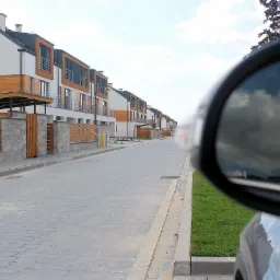 Ulica osiedlowa z szarej kostki brukowej w Ożarowie Mazowieckim, widok z perspektywy kierowcy z lusterkiem bocznym odbijającym chmury.