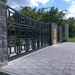 Czarna brama wjazdowa z geometrycznym wzorem, osadzona w kamiennych słupkach, prowadząca na posesję z kostki brukowej, w tle zieleń i budynek mieszkalny.