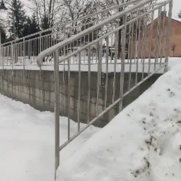 Zewnętrzne schody betonowe z balustradą ze stali nierdzewnej, zasypane śniegiem, w tle budynki i drzewa bez liści.