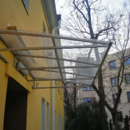 Zadaszenie wejścia do budynku przedszkola, konstrukcja metalowa z przezroczystym wypełnieniem, widok z boku na żółtą elewację i tablicę adresową.