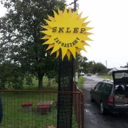 Żółty szyld w kształcie słońca z napisem 'SKLEP ZAPRASZAMY' umieszczony na metalowej konstrukcji, widoczny zza ogrodzenia, w tle drzewa i droga z przejeżdżającymi samochodami w deszczowy dzień.